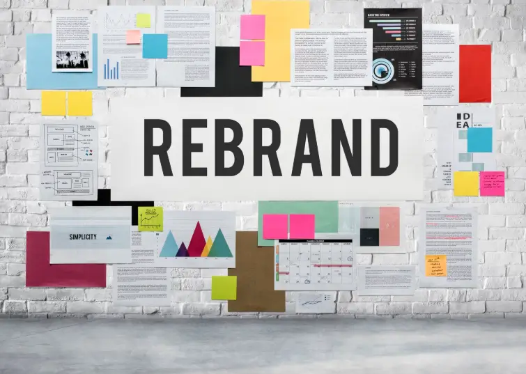 A white brick wall covered in colorful sticky notes, charts, and planning documents surrounds a central "REBRAND" sign, illustrating the organized effort needed to refresh your brand.